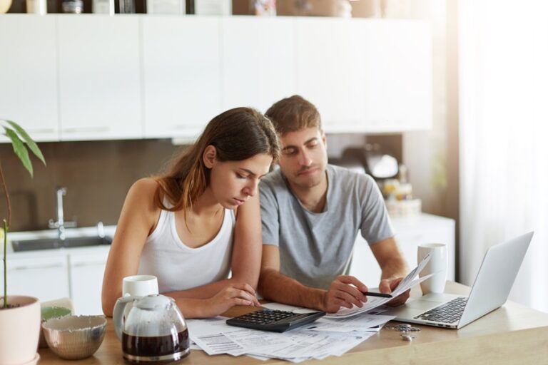 Young couple calculating their family budget, studying documents, using laptop computer and calculator. Female and male sitting in kitchen, analyzing something. Finances and payments concept
