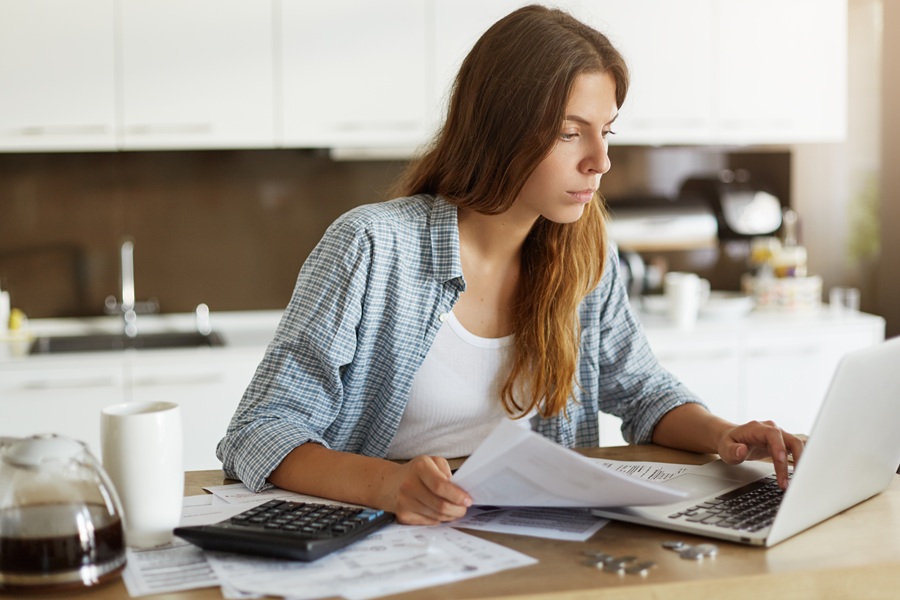 Beautiful young female wearing casual shirt calculating family budget, looking through expenses while managing home accounts, paying utility bills online using wifi on generic notebook computer