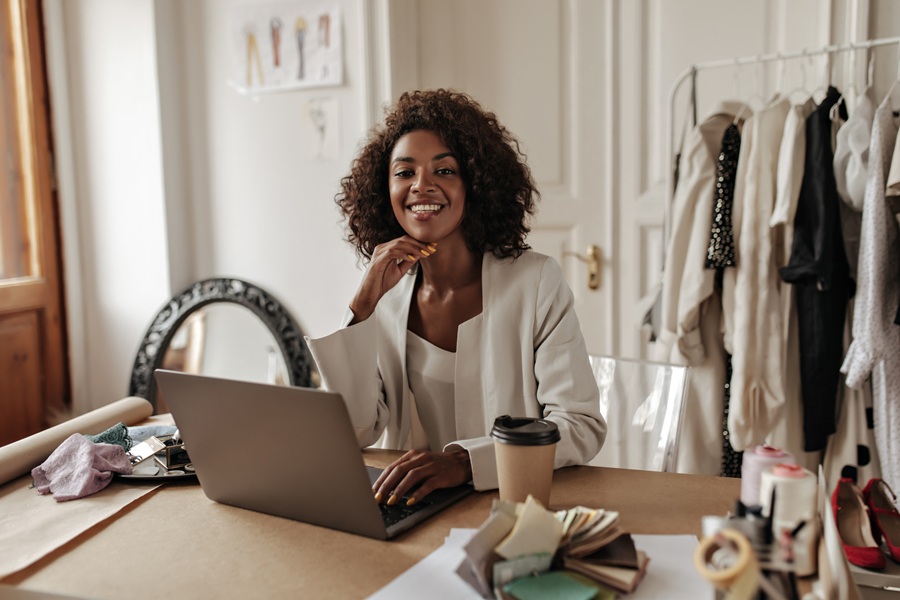 Charming young dark-skinned woman in stylish jacket and blouse smiles, looks at camera, works in laptop and poses in office.